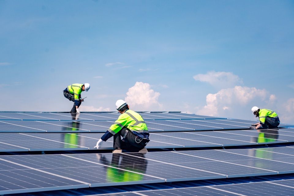 Workers in safety gear installing solar panels on a rooftop, blue sky in background.
