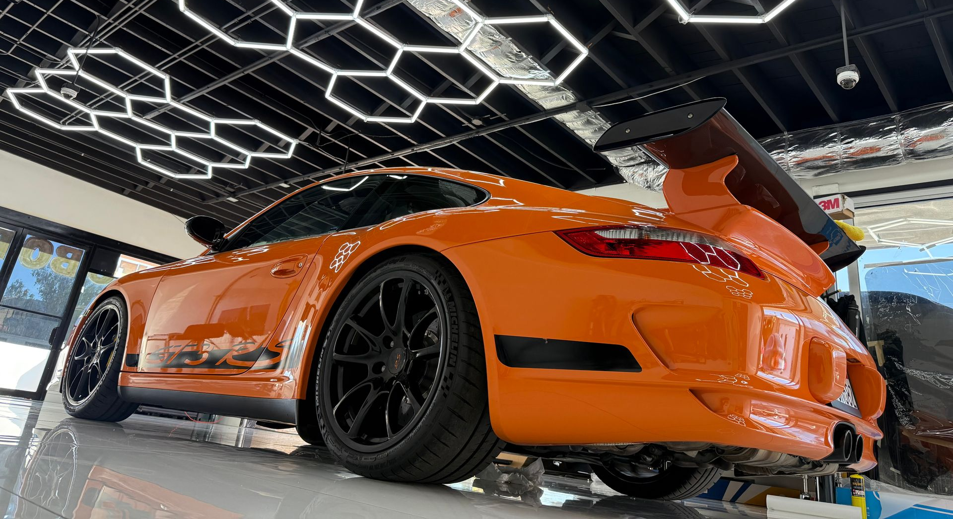 Orange sports car with black wheels displayed indoors under geometric ceiling lights