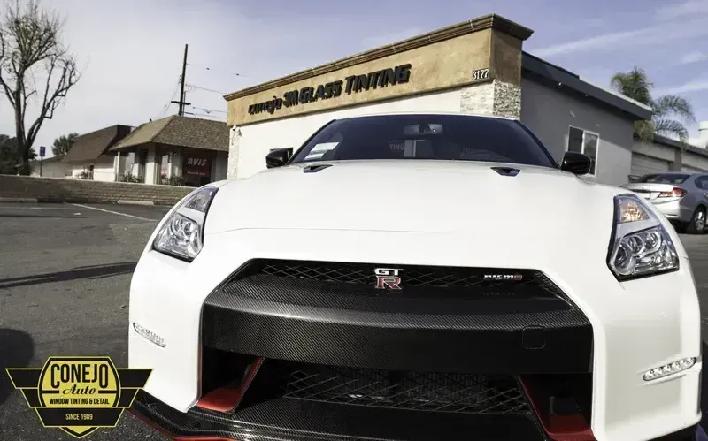 White Nissan GT-R parked outside a shop, front view with a black grille and red tow hook.