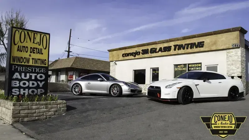 Auto body shop with sign and two parked sports cars outside under a blue sky.