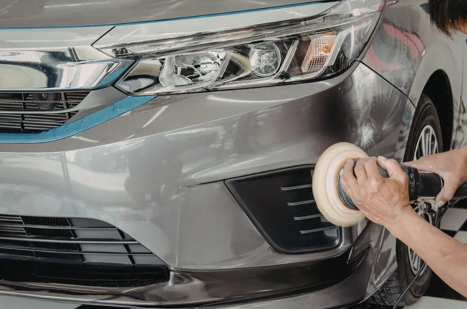 Person polishing the front bumper of a silver car with a buffer in a garage