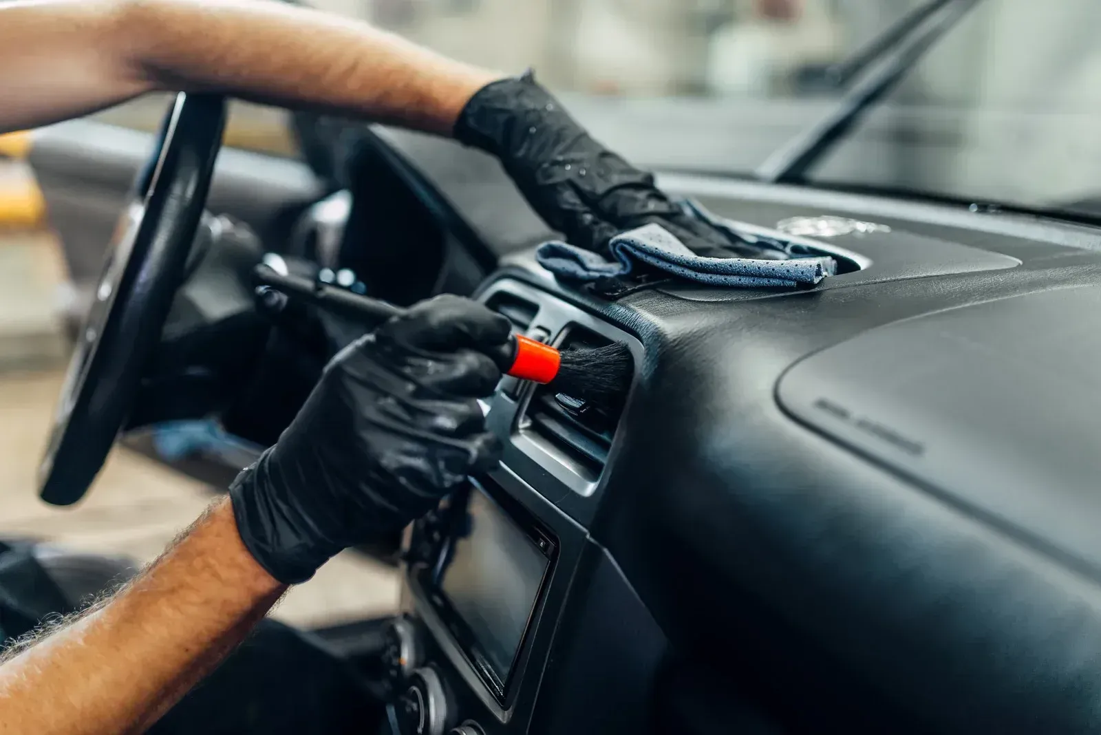 Gloved hands cleaning a car dashboard and steering wheel with a cloth and spray bottle