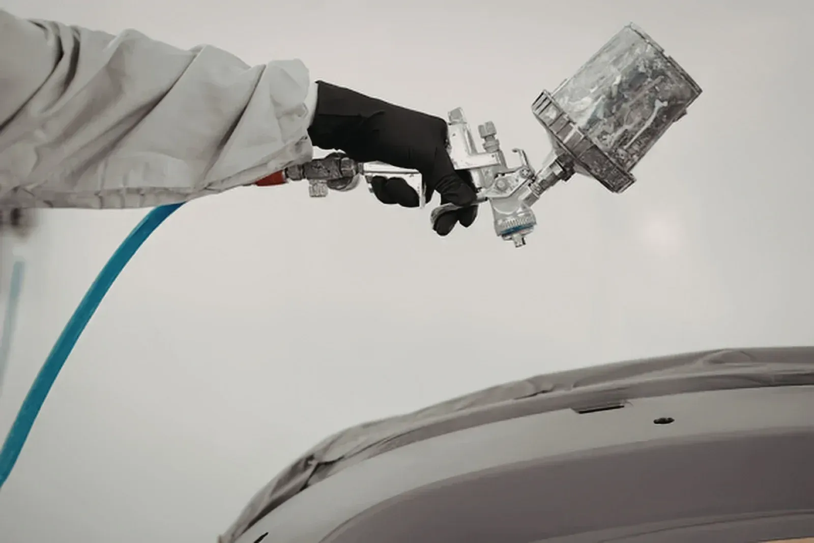 Gloved worker spray-painting a car hood with a paint gun and blue hose