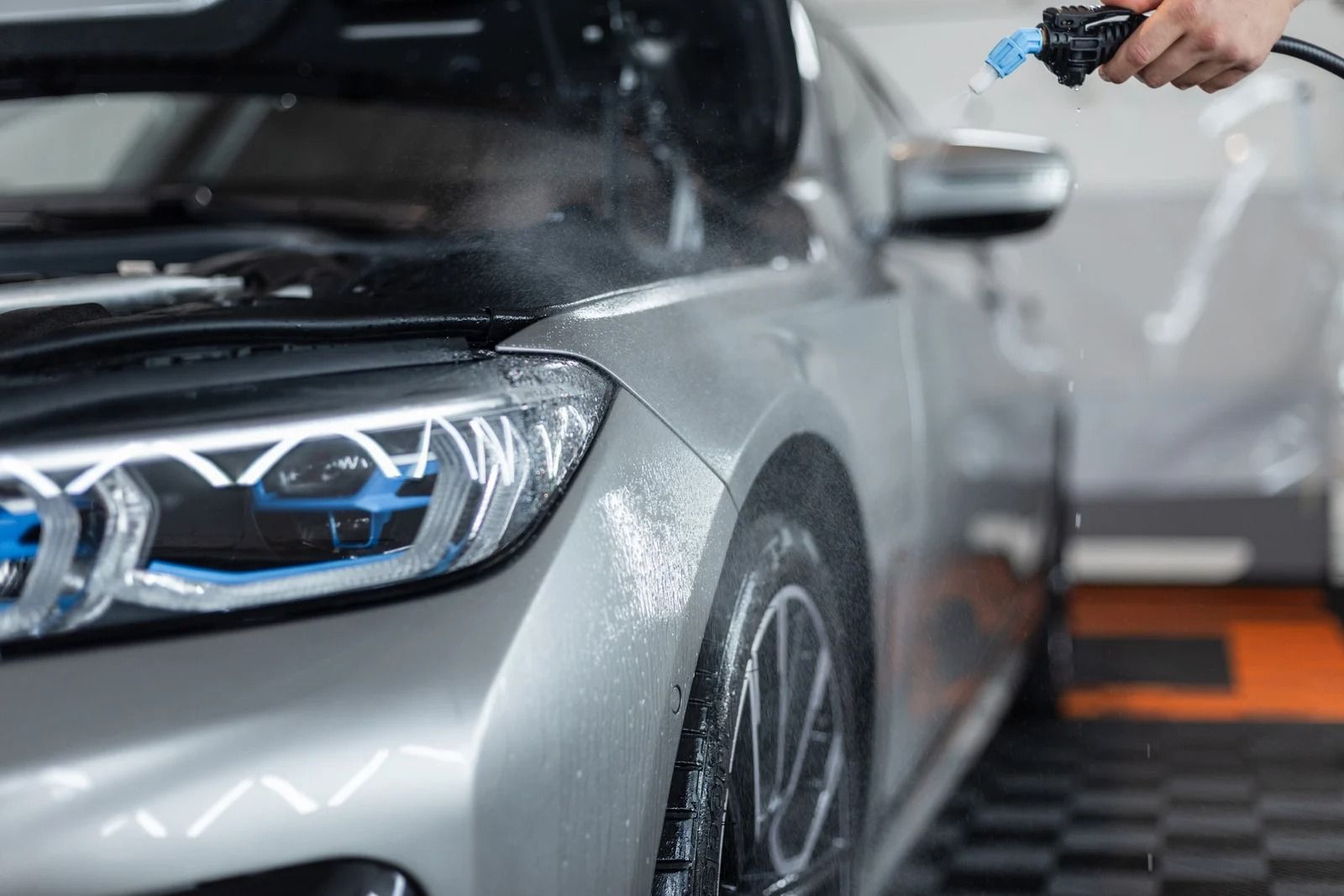 Silver car being sprayed with water in a car wash, focusing on the front headlight and wheel