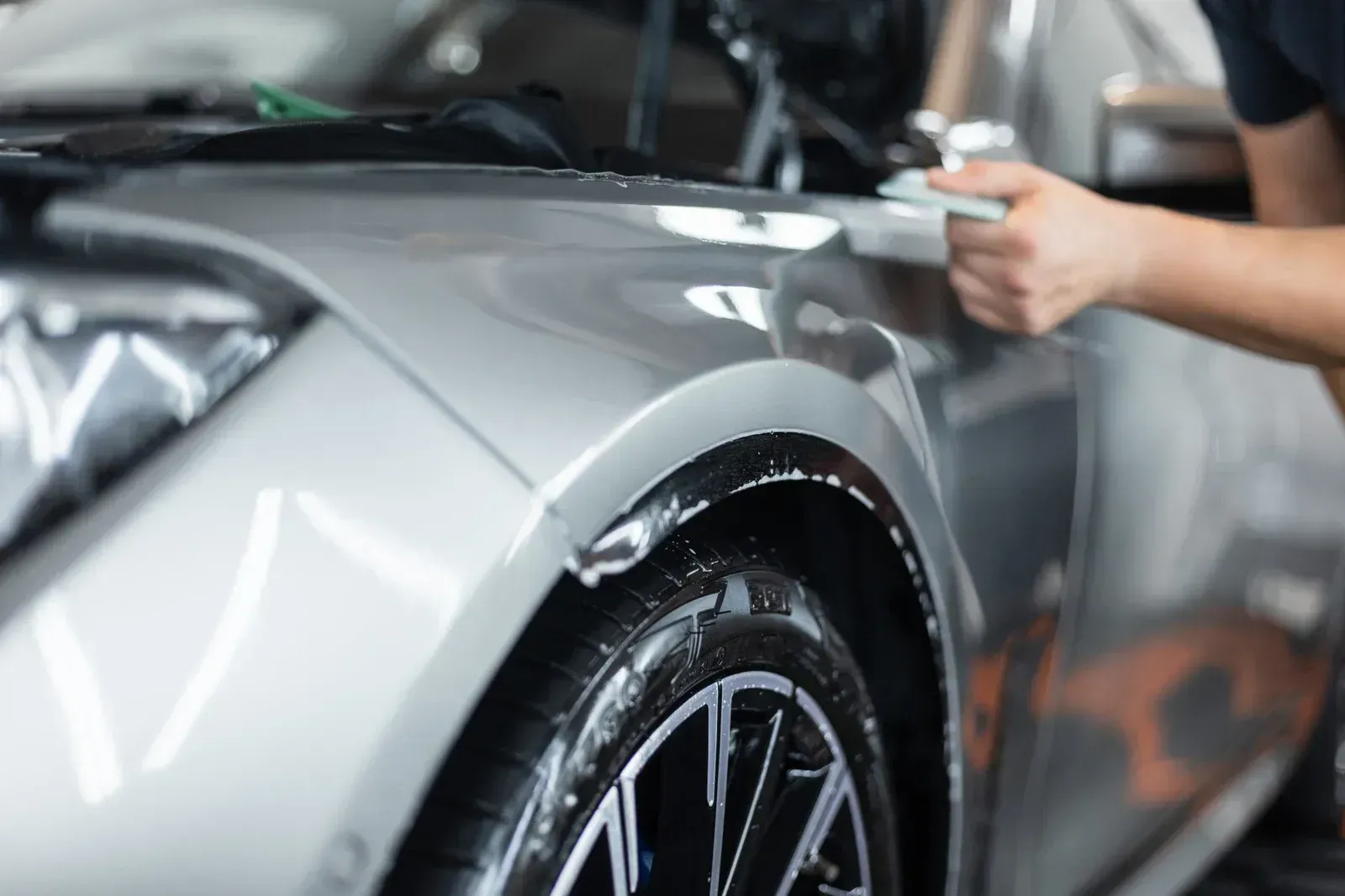 Person polishing a silver sports car’s front fender in an auto detailing shop
