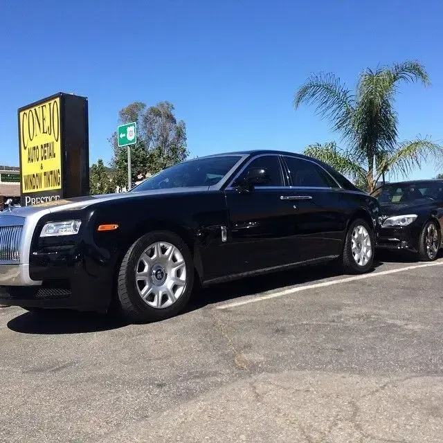 Black luxury sedan parked in a sunny lot beside a yellow roadside sign and palm trees.
