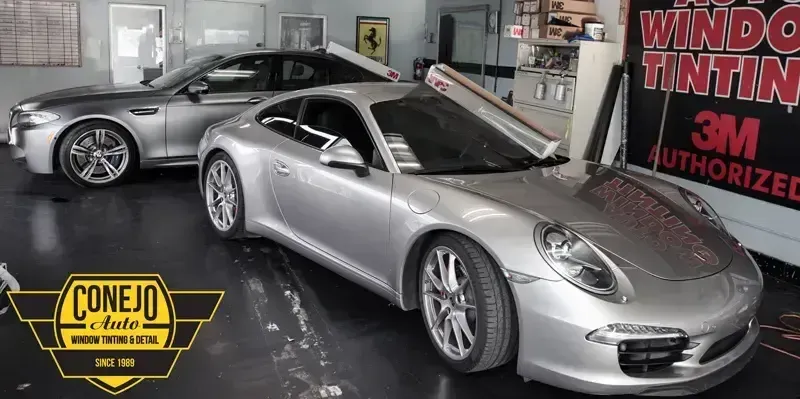 Two silver sports cars inside a window tinting shop, with a “Window Tinting 3M Authorized” sign visible.