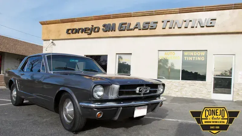 Black classic Ford Mustang parked outside a glass tinting shop on a sunny day.