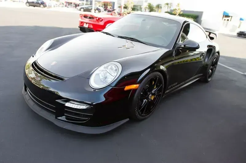 Black Porsche sports car parked outdoors, front-left view with red car in the background