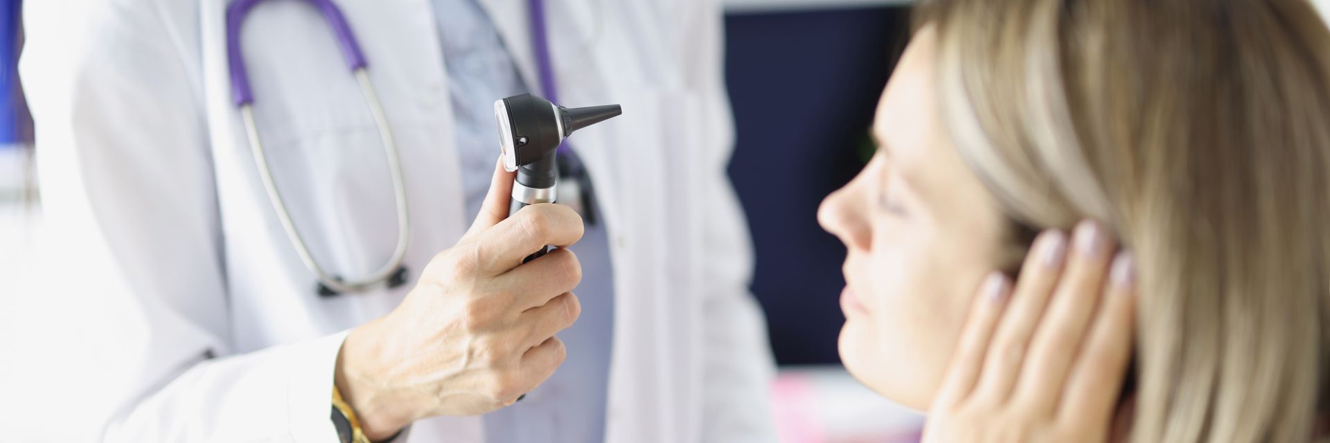 An audiologist holds an otoscope while examining a seated patient’s ear in a clinical exam room