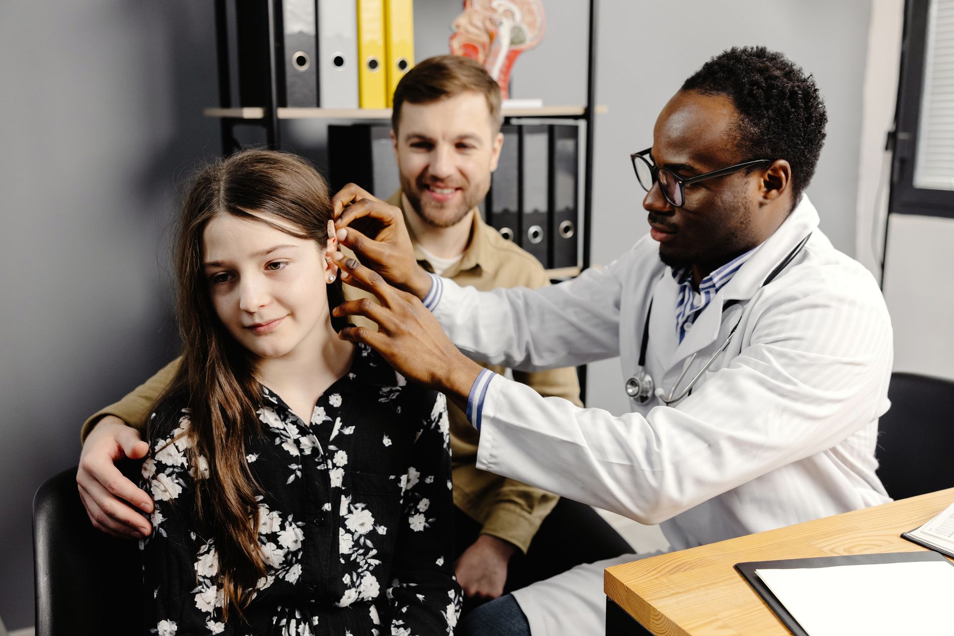 A doctor is inserting a hearing aid into a young girl's ear in his office while a man is watching. A doctor is inserting a hearing aid into a young girl's ear in his office while a man is watching.