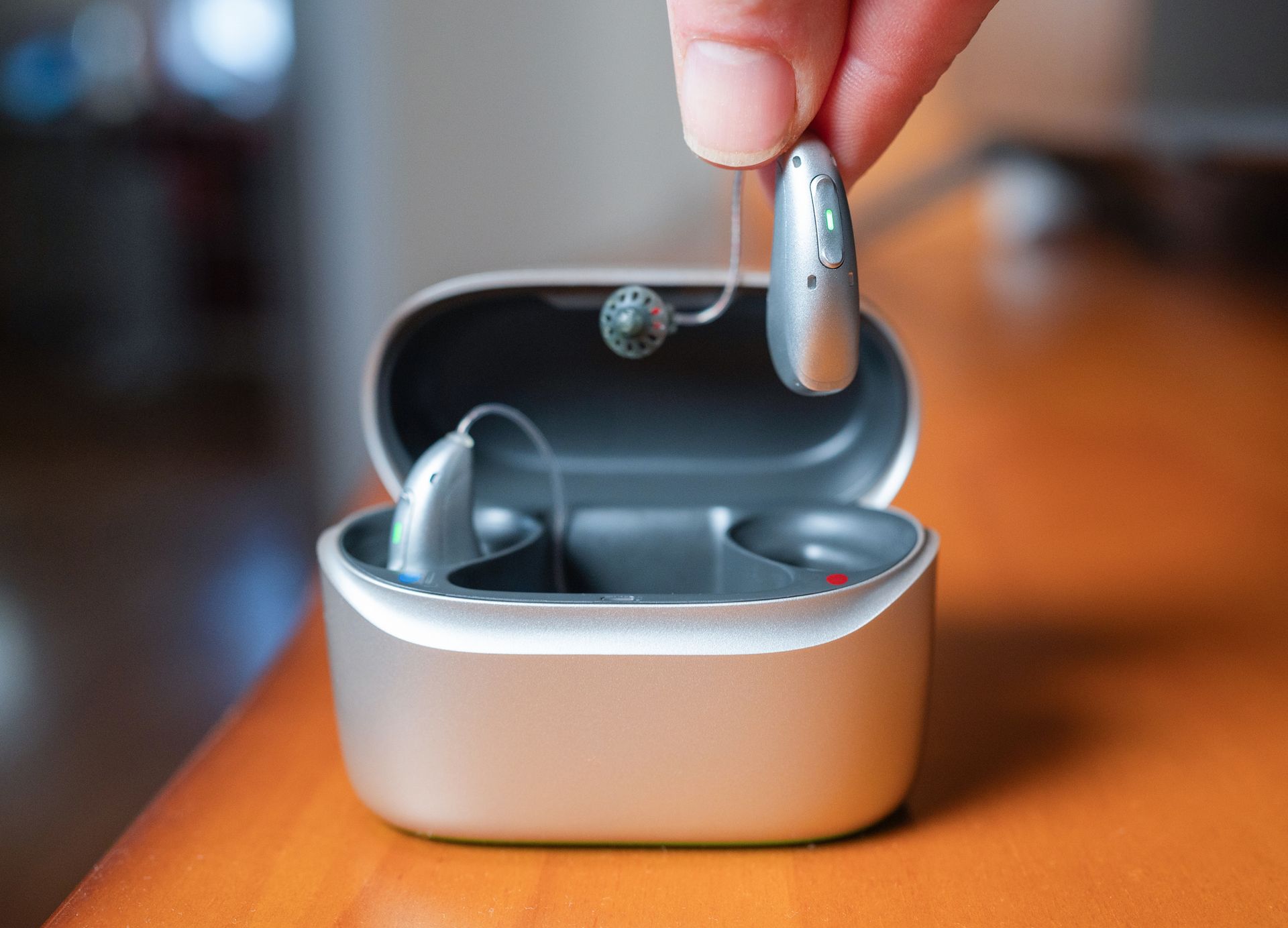 Hand holding hearing aid above a charging case containing two other hearing aids on a wooden table.
