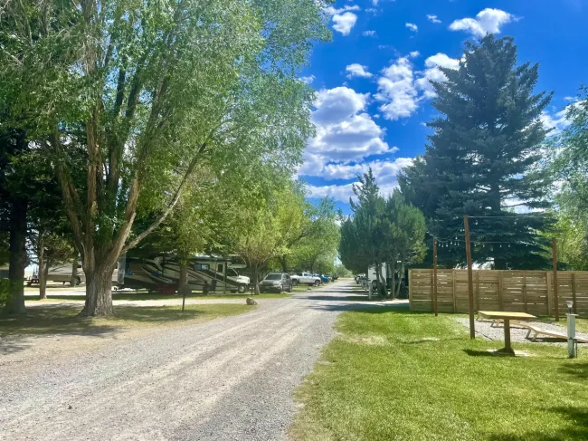 Gravel road through a campground with trees, RVs, picnic table, and blue sky.