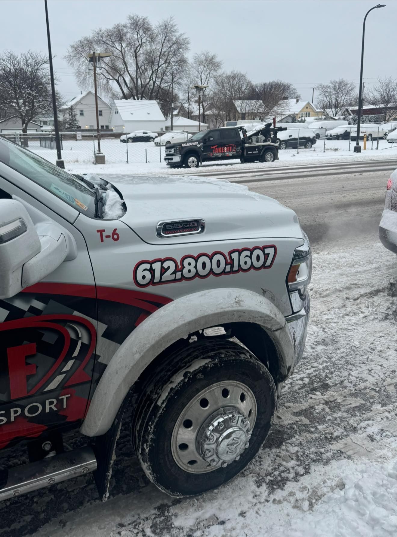 Tow truck with phone number on side in snowy setting; another tow truck in the background.