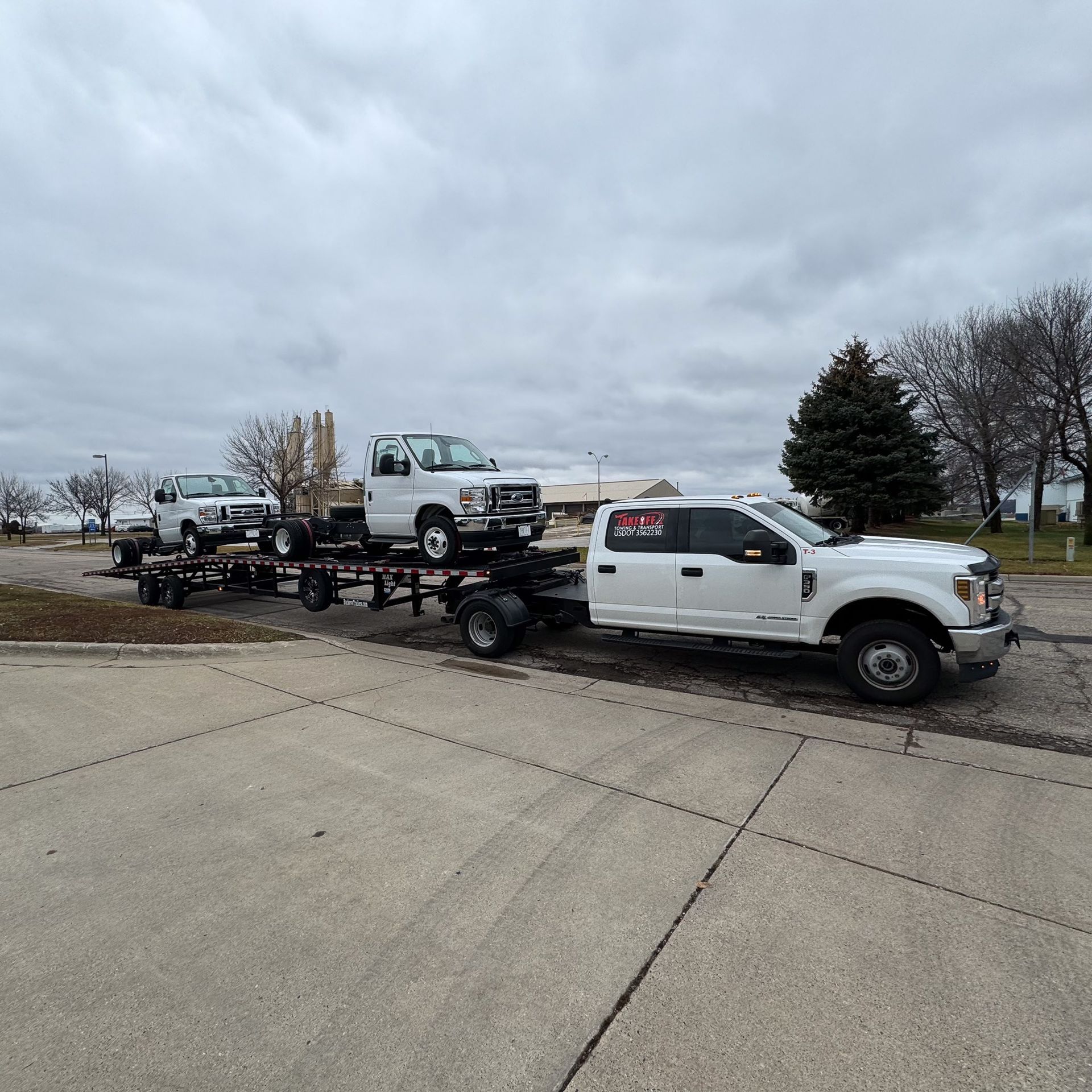 White truck towing a trailer with two white trucks on a gray concrete surface. Overcast sky.