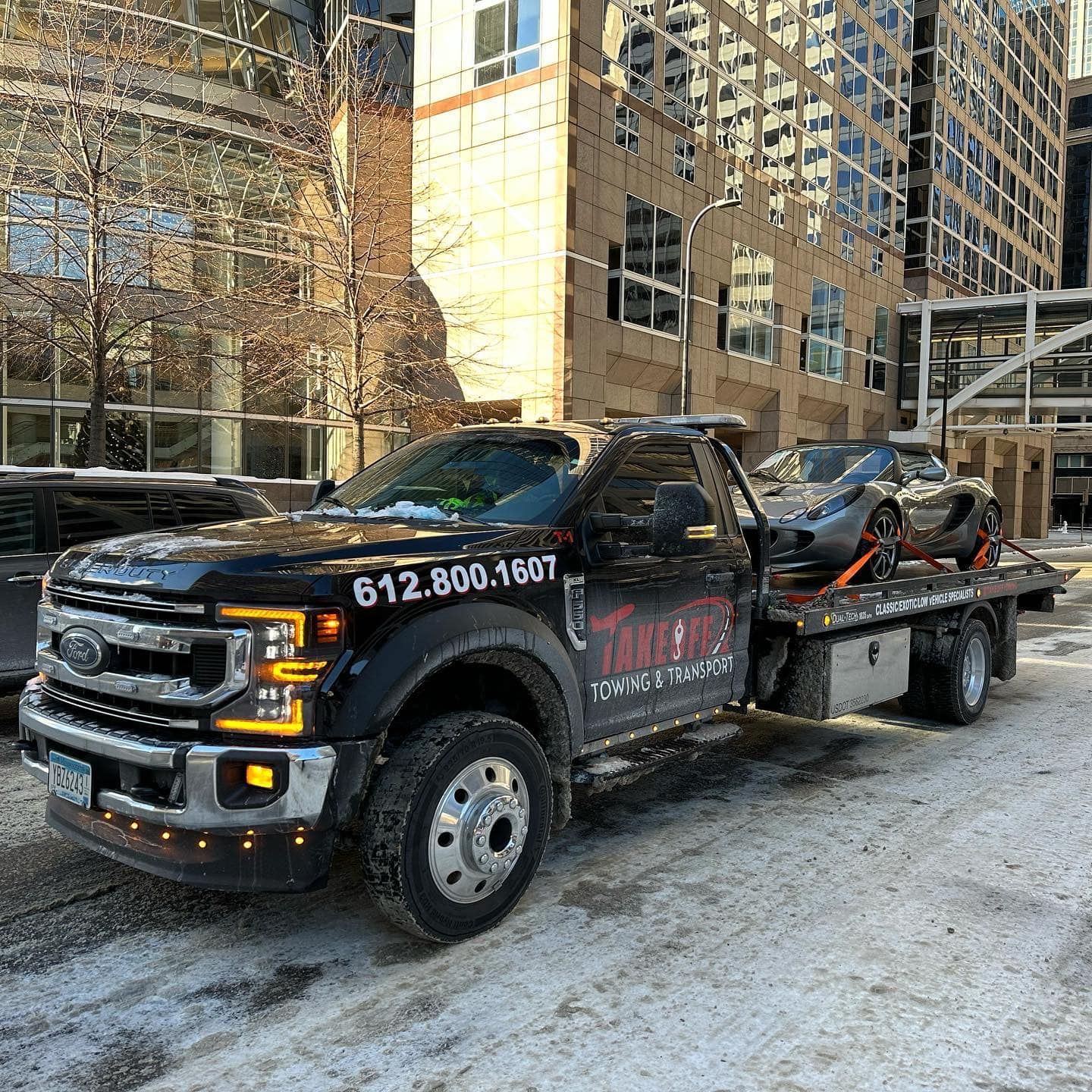 Black tow truck with a grey car on its flatbed, in a city setting.