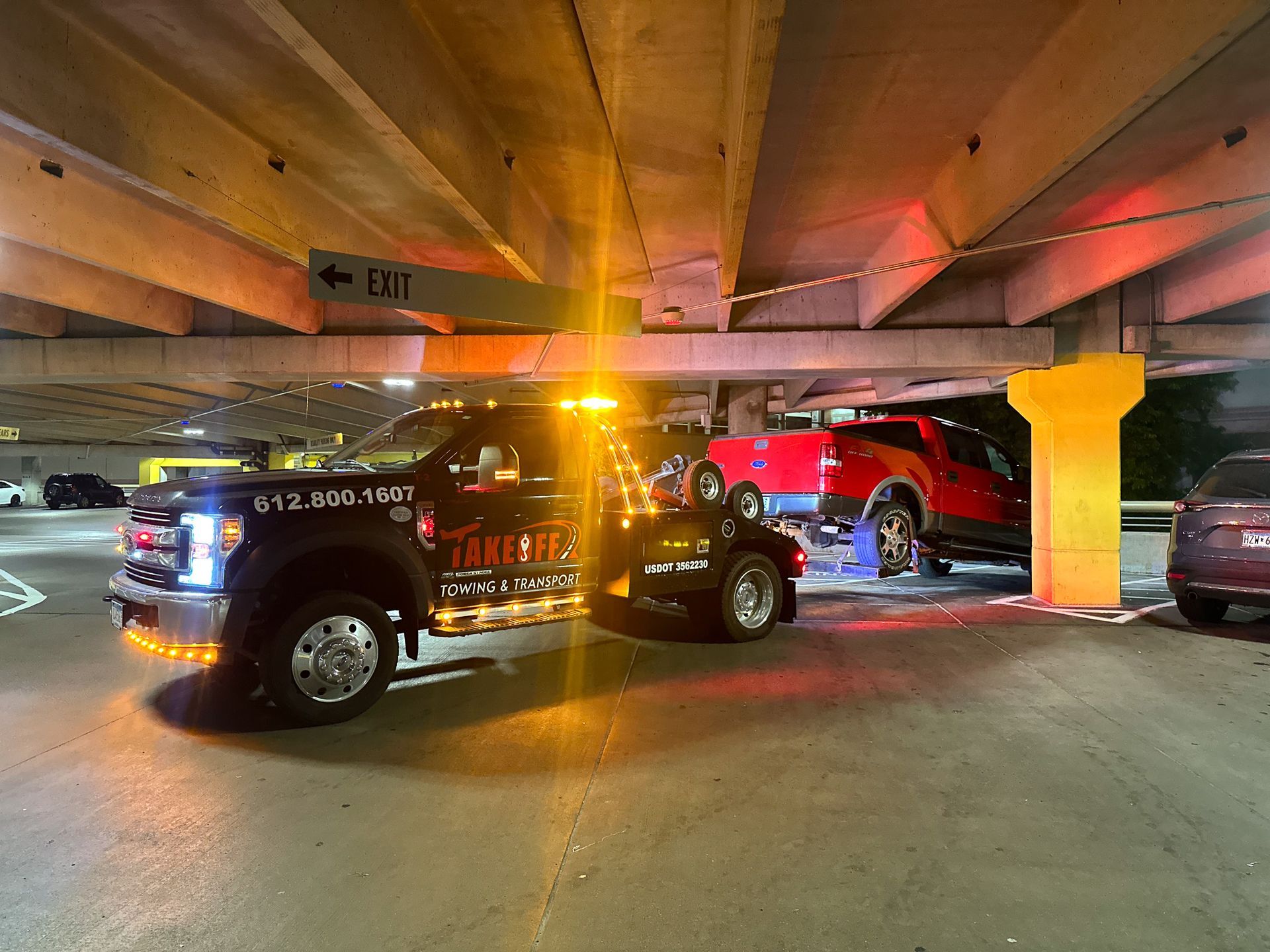 Tow truck under a parking garage, towing a red pickup truck with flashing amber lights.