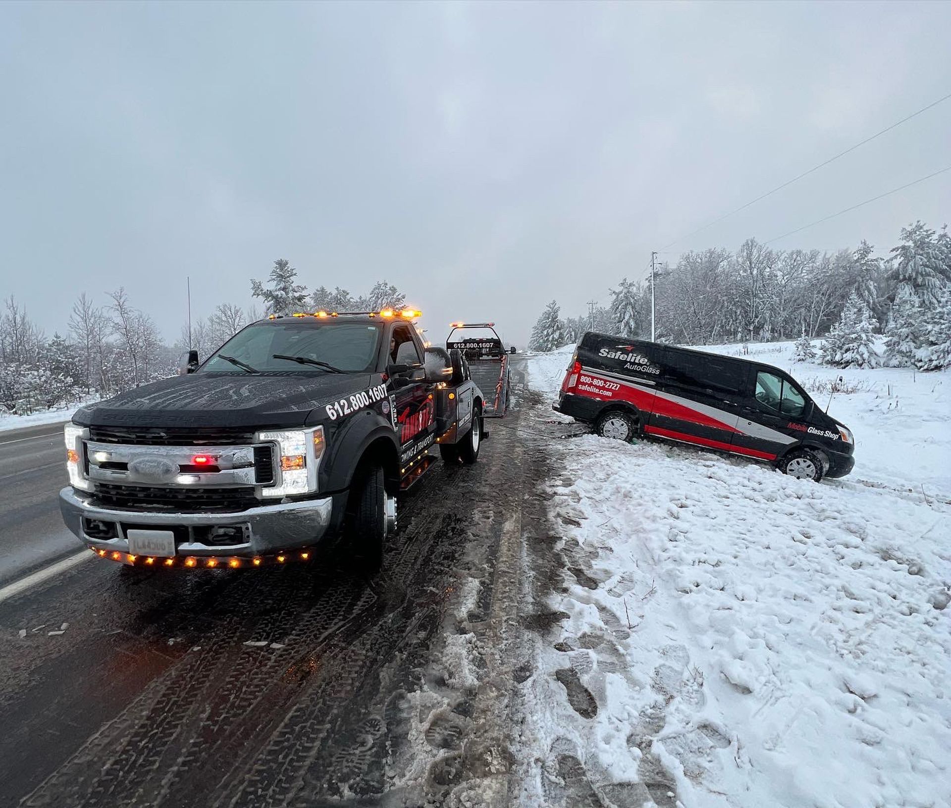 Tow truck pulling a black and red van from a snow-covered roadside. Snowy day, dark truck.