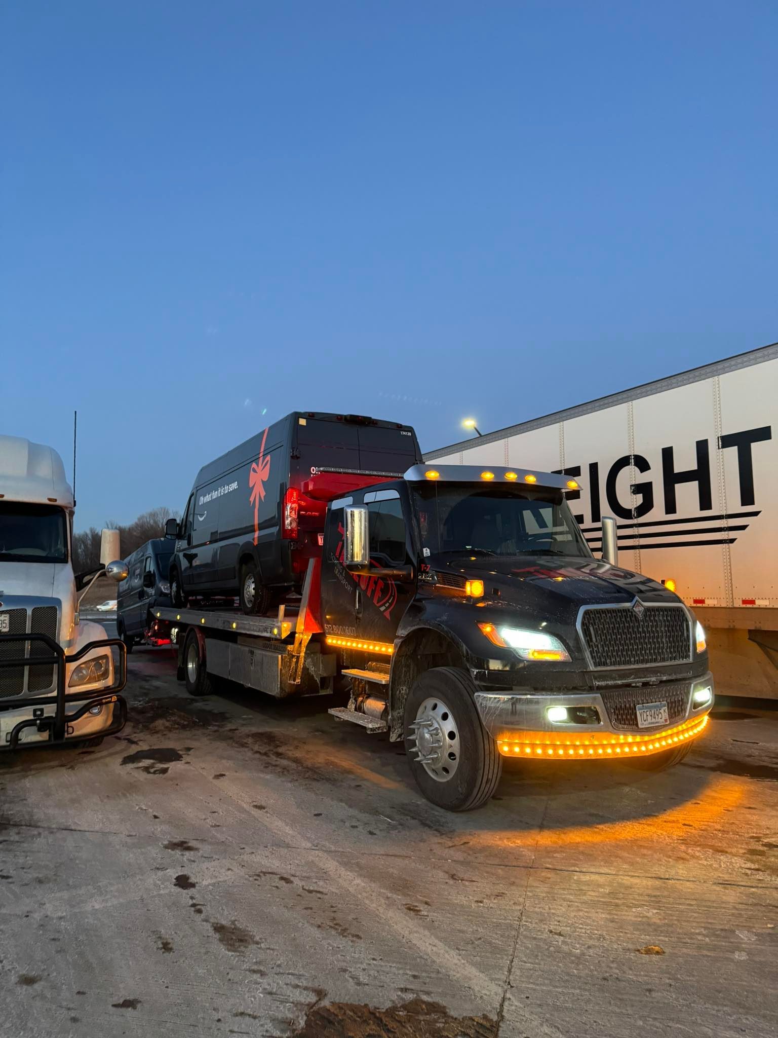 Black tow truck hauling a black vehicle, parked next to a white freight truck at dusk.