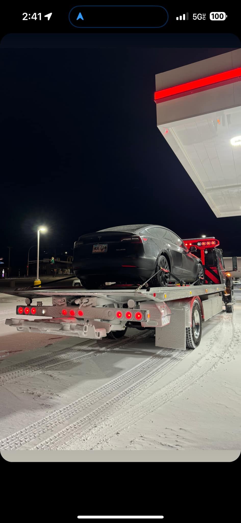 A black car is on a flatbed tow truck at a gas station at night in winter.