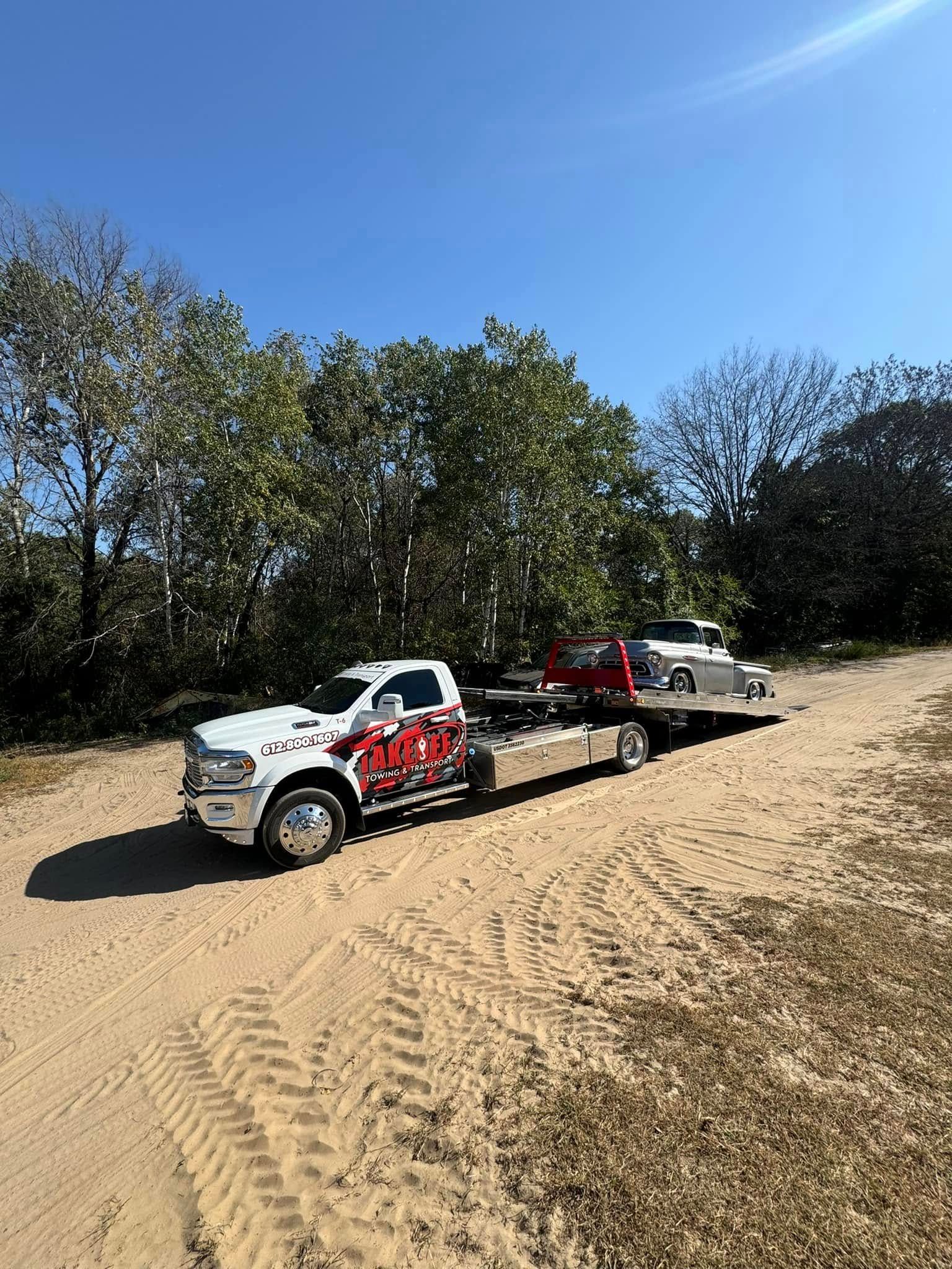 Tow truck with a car on its flatbed on a dirt road, trees in background, blue sky.