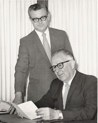 Two men in suits, one seated, reading a document. The other stands behind, looking on. Both wear glasses.