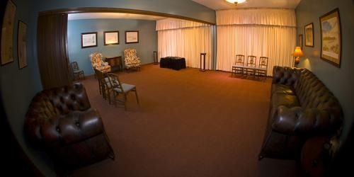 A room with dark furniture and brown carpet, curtains, chairs, and pictures on the wall.