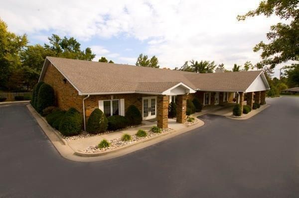Tan brick building with a brown roof and covered walkway; surrounded by bushes and a paved driveway.
