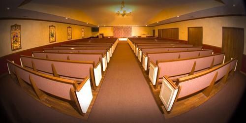 Interior of a church or auditorium, with rows of wooden pews facing a stage.