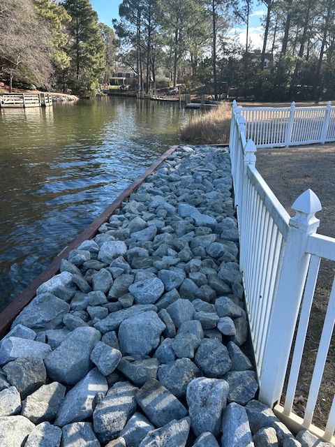A rocky shoreline with a white fence along the grass and a lake in the background.