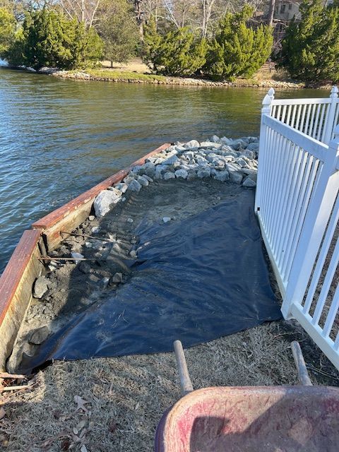 Shoreline construction with dark fabric, rocks, and white fence. Water and trees in the background.