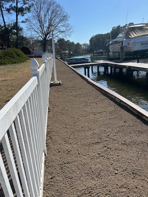 A waterside walkway with white picket fence, brown ground, and docks with boats on the water.