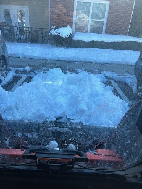 View from inside a vehicle; a snow-filled plow in front, clearing a snowy parking spot.