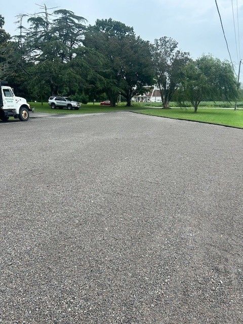 Gravel parking lot with a truck, cars, and trees in the background. Green grass and a house visible.