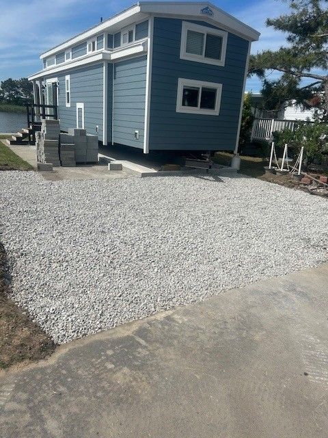 Gravel driveway under construction, bordered by grass, trees, and a row of evergreen trees, on a sunny day.