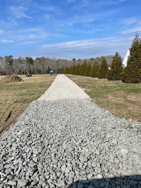 Gravel driveway in progress, with grass on either side, trees in the background, and a blue sky.