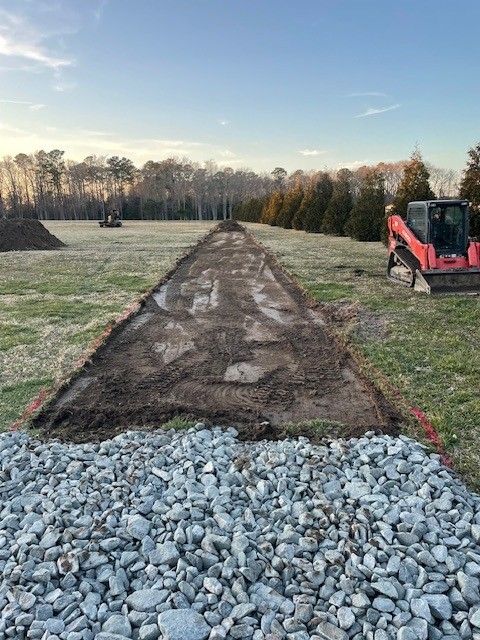 Gravel pile and cleared earth path for construction; small red skid steer sits on the right.