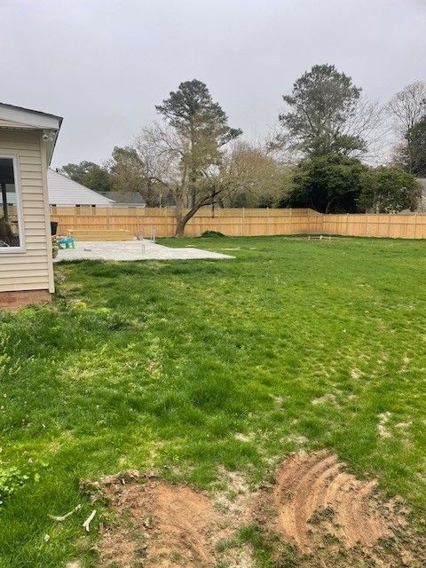 Grassy backyard with wooden fence and concrete slab, overcast sky, and part of a house.