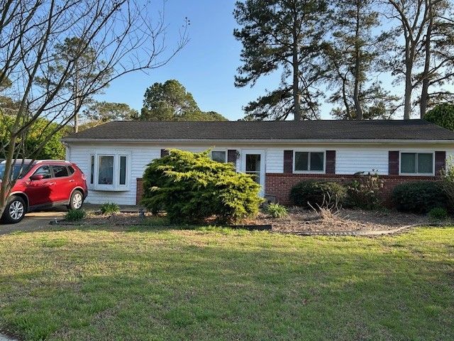 A single-story house with white siding, red shutters, and a brick facade, set in a grassy yard.