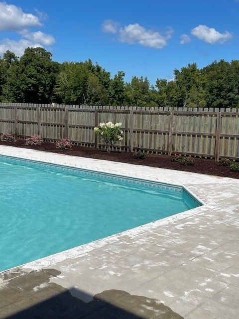 Poolside view with blue water, concrete deck, brown fence, and cloudy sky.