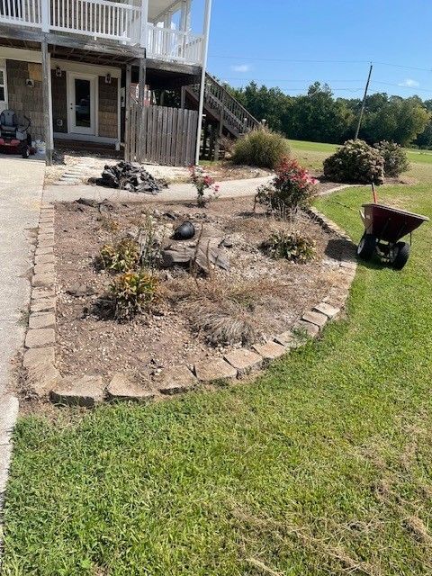 A flower bed edged with stone blocks, with shrubs and a wheelbarrow, next to a lawn and a house.