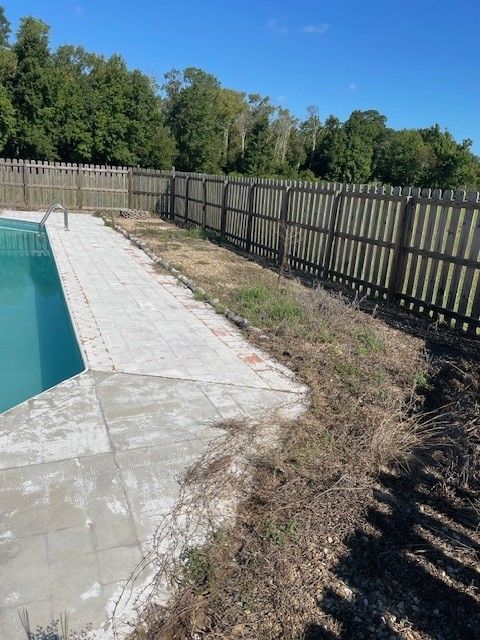 Poolside area with a fence, overgrown grass, and trees under a blue sky.