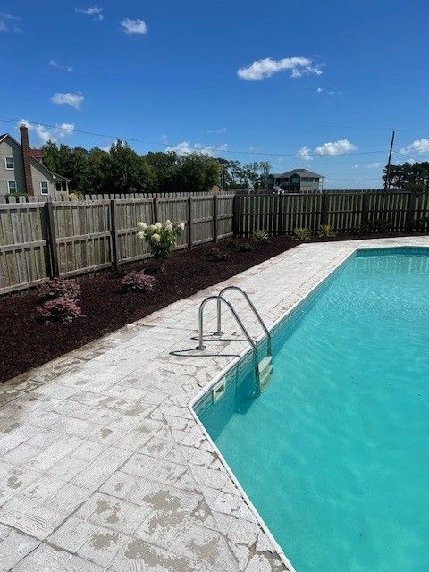 Poolside view with blue water, white paving, wooden fence, and garden beds under a bright blue sky.