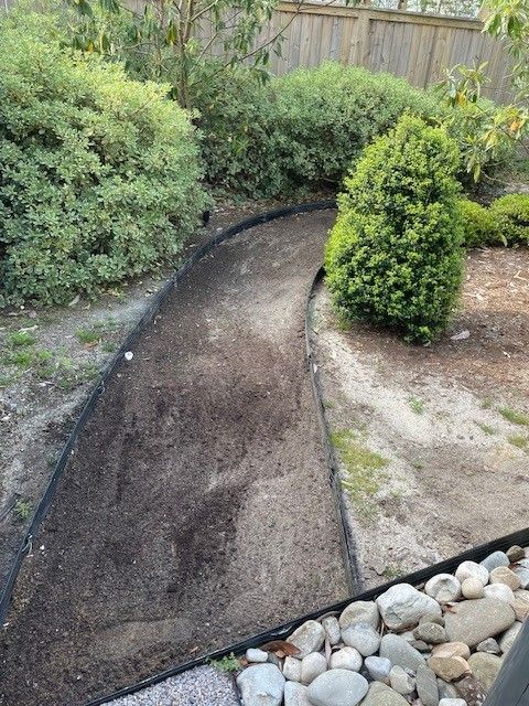 A winding garden path of dark soil bordered by black edging, surrounded by greenery and rocks.