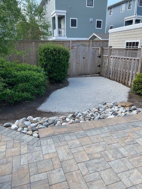 Backyard with patio, gravel area, rocks, gate, and green shrubs. Houses visible in the background.