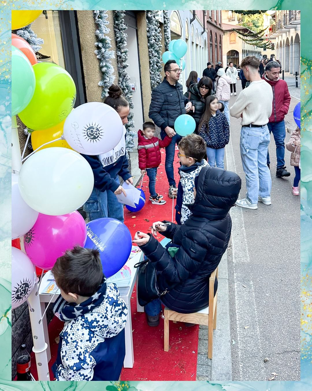 Persone radunate all'aperto; palloncini e bambini vicino a un tavolo. Alcuni osservano una persona con una giacca con cappuccio.