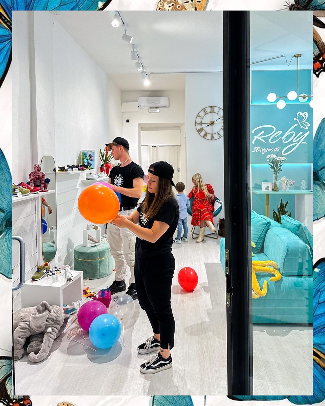 Persone in negozio con palloncini. Scaffali con giocattoli, divano verde acqua, insegna sul muro e cornice decorativa a forma di farfalla.