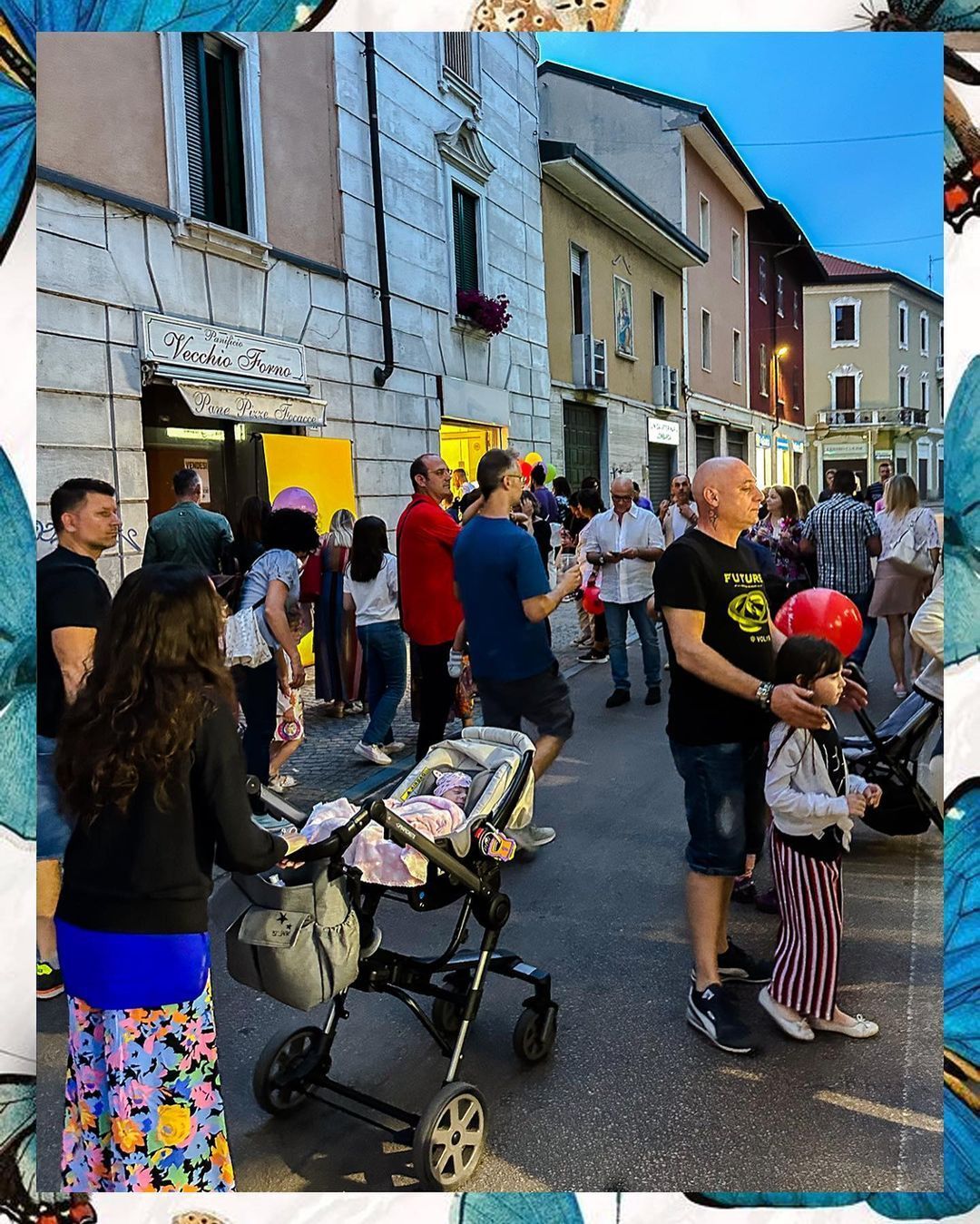 La gente si raduna in una strada con degli edifici, alcuni guardano uno spettacolo, con un passeggino in primo piano.