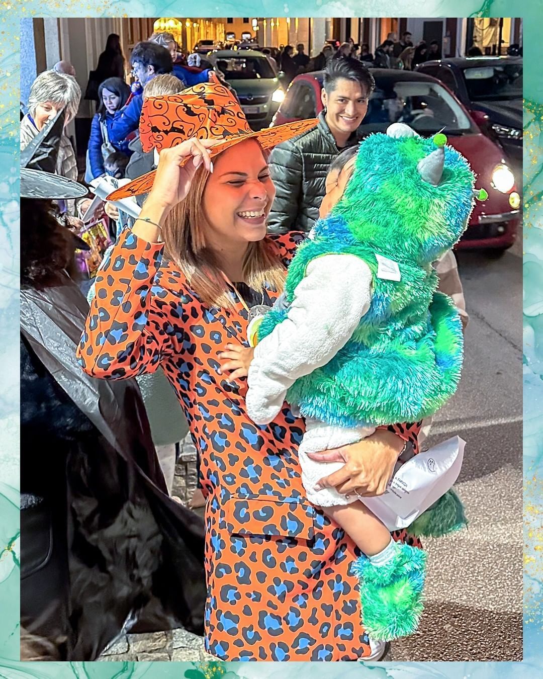 Una donna vestita di arancione tiene in braccio un bambino vestito da mostro; sorridono in una strada di notte.