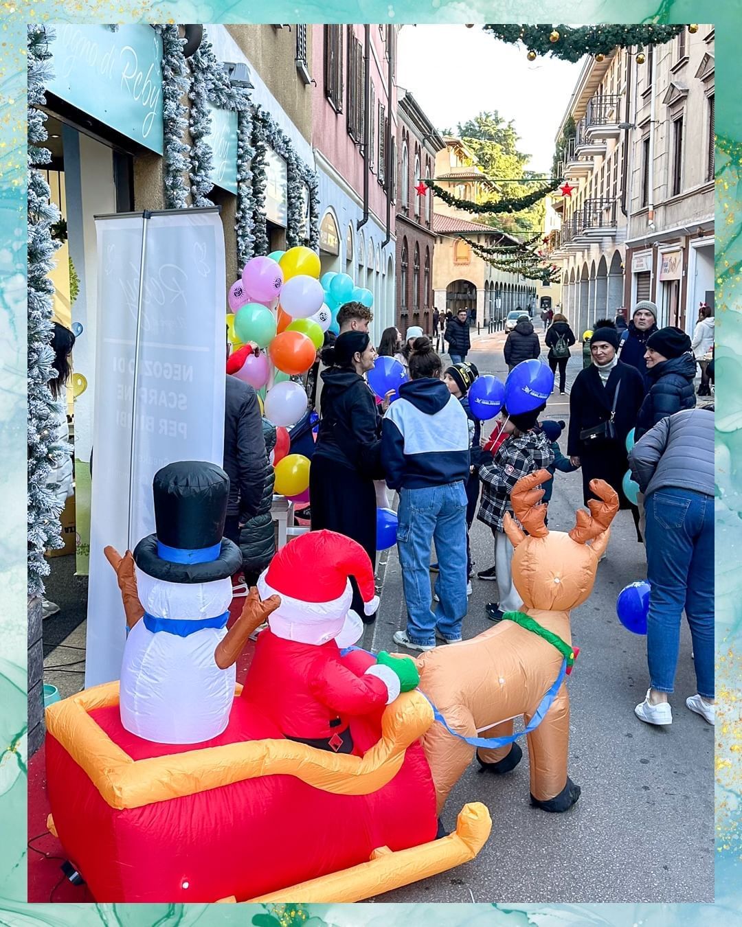 Scena di strada festosa con gonfiabili raffiguranti Babbo Natale e renne, persone, palloncini e decorazioni natalizie.